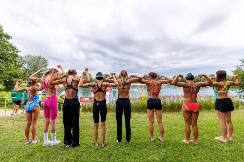 Gruppe junger Schwimmer zeigt ihre Muskeln, Rücken zur Kamera, mit Startnummern auf den Schultern am Gewässer.