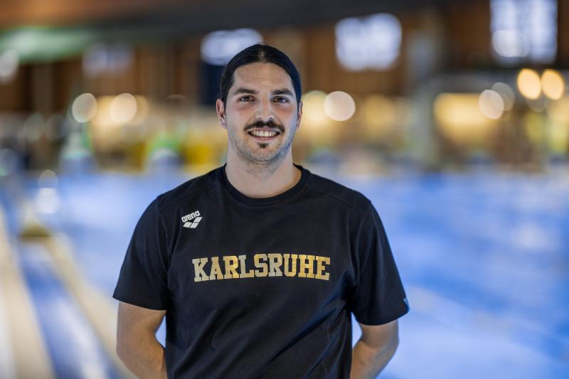 Mann mit dunklem Haar und Schnurrbart trägt ein schwarzes T-Shirt mit dem Aufdruck „Karlsruhe“ vor einem Schwimmbecken.