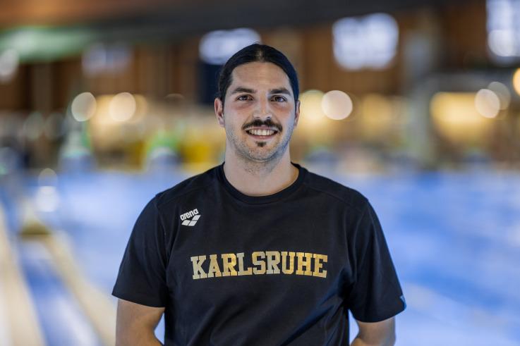 Mann mit dunklem Haar und Schnurrbart trägt ein schwarzes T-Shirt mit dem Aufdruck „Karlsruhe“ vor einem Schwimmbecken.