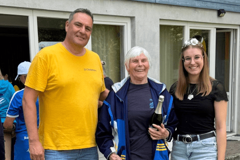 Drei Personen stehen vor einem Gebäude, eine hält einen Pokal, während die anderen sie anlächeln. Hintergrund zeigt eine Gruppe.