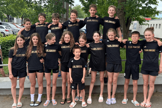 Gruppe von 15 Jugendlichen in schwarzen T-Shirts mit der Aufschrift „Karlsruhe“ steht vor einer grünen Kulisse.