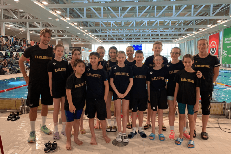 Gruppe von Kindern und Trainern in schwarzen T-Shirts mit der Aufschrift „Karlsruhe“ am Schwimmbecken.
