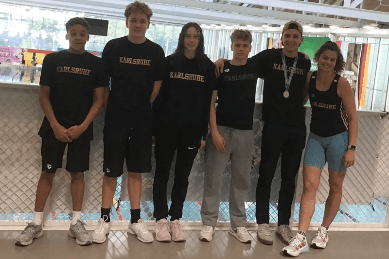 Sechs Jugendliche mit Sportbekleidung stehen lächelnd vor einer Schwimmhalle, einige tragen Medaillen.