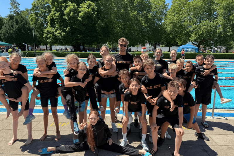 Eine Gruppe von Kindern und Jugendlichen in schwarzen T-Shirts mit „Karlsruhe“ posiert lächelnd am Schwimmbecken.