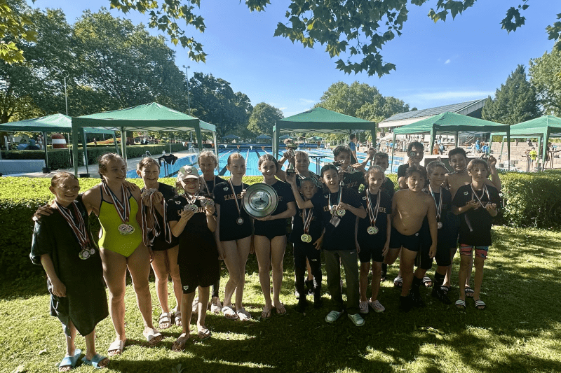 Gruppenshot von Jugendlichen in Badekleidung mit Medaillen und Pokal auf einer Wiese neben einem Schwimmbad.