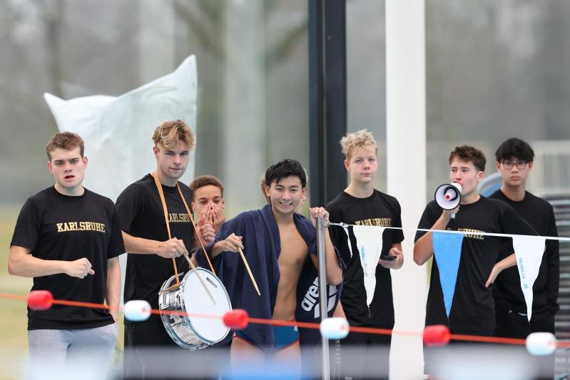 Gruppe junger Schwimmer in schwarz-gelben T-Shirts, dabei ein Trommelspieler und ein Megafonträger am Beckenrand.