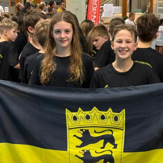 Zwei Kinder halten eine schwarze und gelbe Flagge mit einem Wappen und stehen vor einer Gruppe von Menschen.