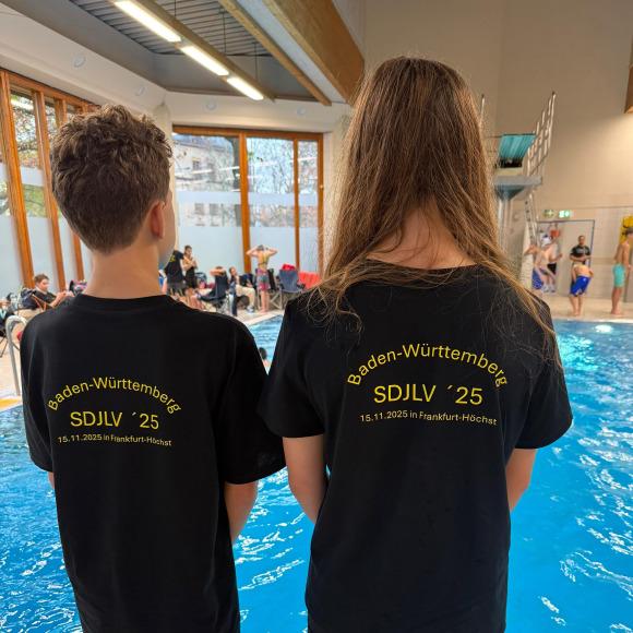 Zwei Kinder stehen mit Rücken zur Kamera am Beckenrand, tragen schwarze T-Shirts mit Aufdruck zu einem Schwimm-Event.