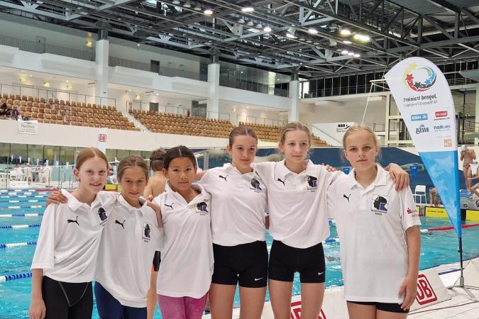 Sechs junge Schwimmerinnen in einheitlichen T-Shirts stehen am Schwimmbeckenrand einer modernen Schwimmhalle.
