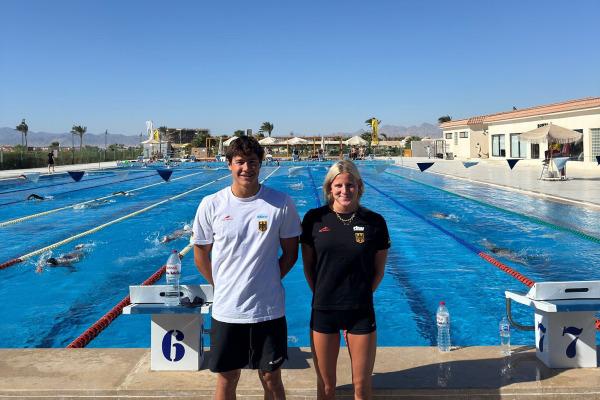 Zwei Schwimmer stehen vor einem Schwimmbecken, umgeben von Schwimmbahnen und palmenbestandenen Landschaften.