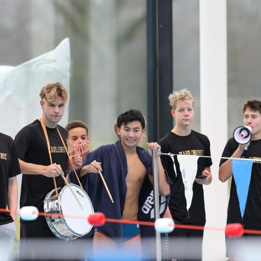 Gruppe junger Schwimmer in schwarz-gelben T-Shirts, dabei ein Trommelspieler und ein Megafonträger am Beckenrand.