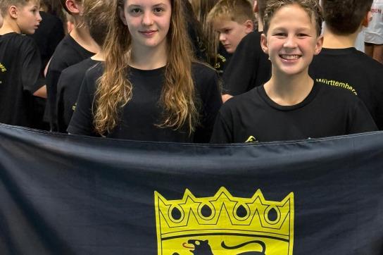 Zwei Kinder halten eine schwarze und gelbe Flagge mit einem Wappen und stehen vor einer Gruppe von Menschen.