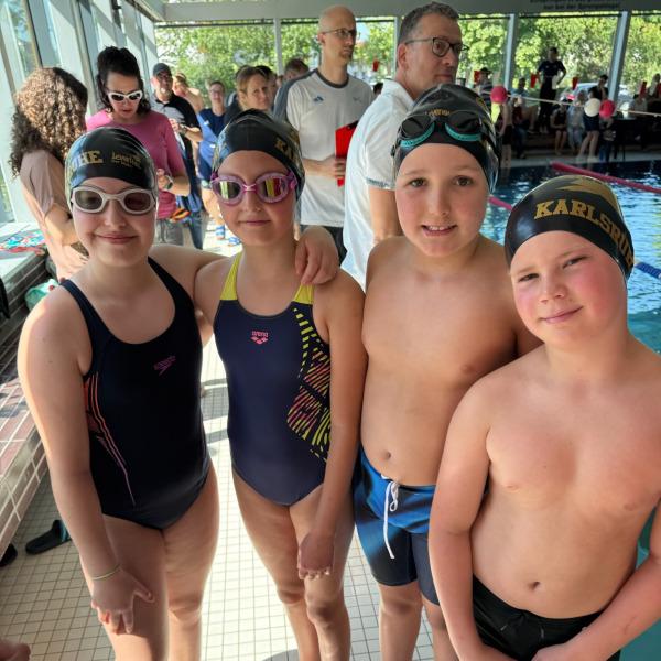 Vier Kinder in Schwimmbekleidung und Badekappen stehen am Rand eines Schwimmbeckens. Zuschauer sind im Hintergrund sichtbar.