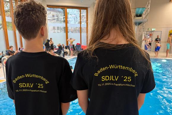 Zwei Kinder stehen mit Rücken zur Kamera am Beckenrand, tragen schwarze T-Shirts mit Aufdruck zu einem Schwimm-Event.