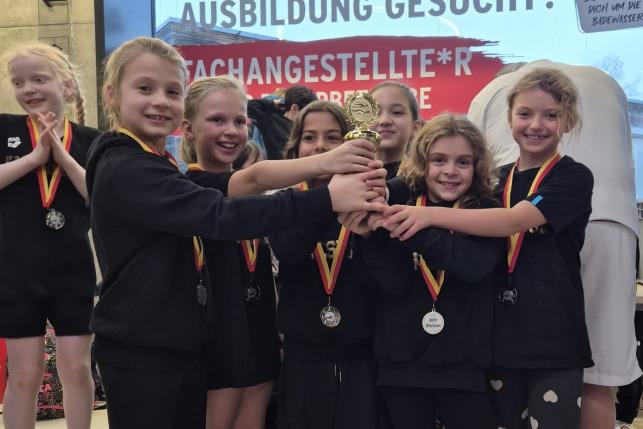 Gruppe von sieben Mädchen in Schwarz mit Medaillen und einem Pokal, stehend vor einem Plakat mit Werbung.