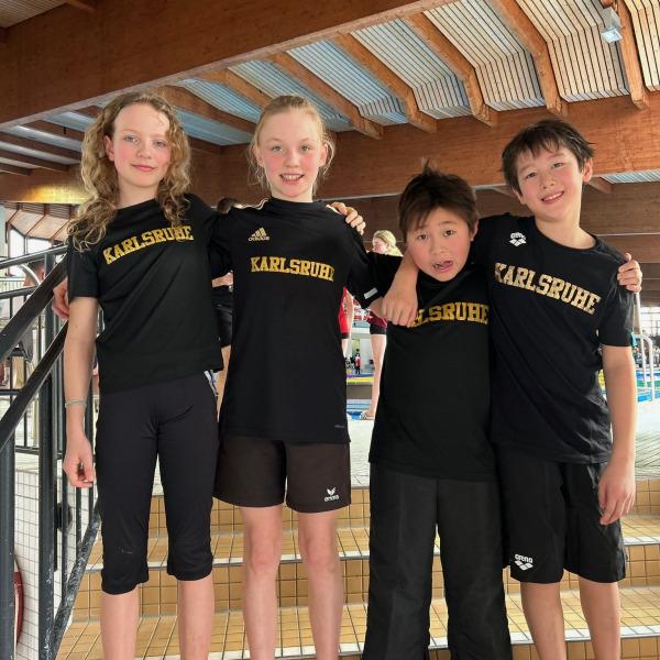 Vier Kinder in schwarzen T-Shirts mit der Aufschrift "Karlsruhe" stehen lachend auf einer Treppe in einem Schwimmbad.