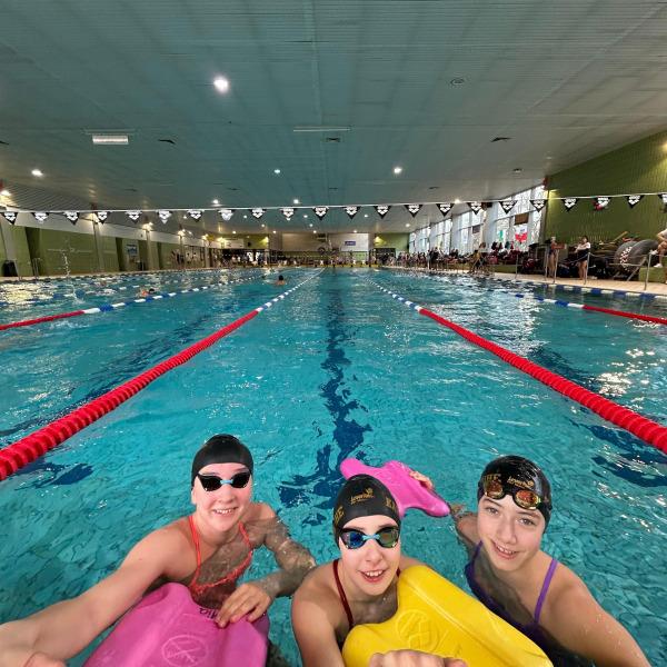 Drei schwimmende Kinder mit Schwimmbrettern posieren am Beckenrand eines Hallenbades.