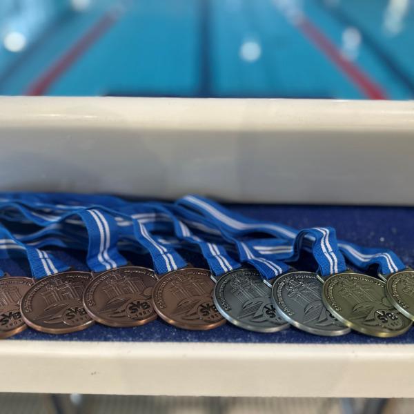 Acht Schwimmmedaillen in Bronze, Silber und Gold hängen an blauen Bändern auf einer Tischkante, dahinter ein Schwimmbecken.