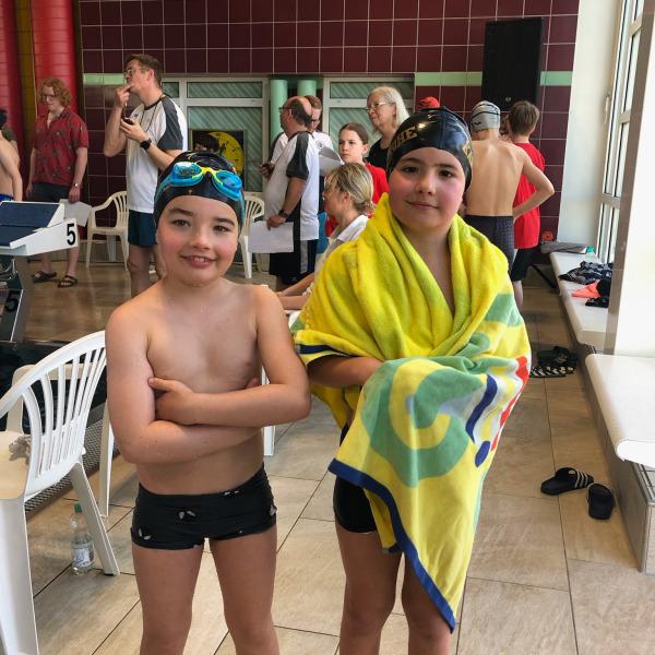 Zwei Kinder in Badesachen stehen in einer Schwimmhalle, eines mit Schwimmbrille, das andere in ein Handtuch gehüllt.