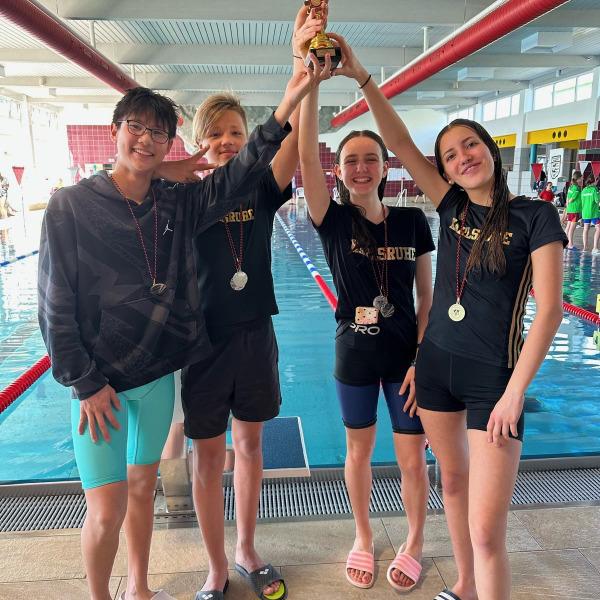 Vier Schwimmer stehen am Schwimmbecken und halten gemeinsam einen Pokal hoch, alle tragen Medaillen.