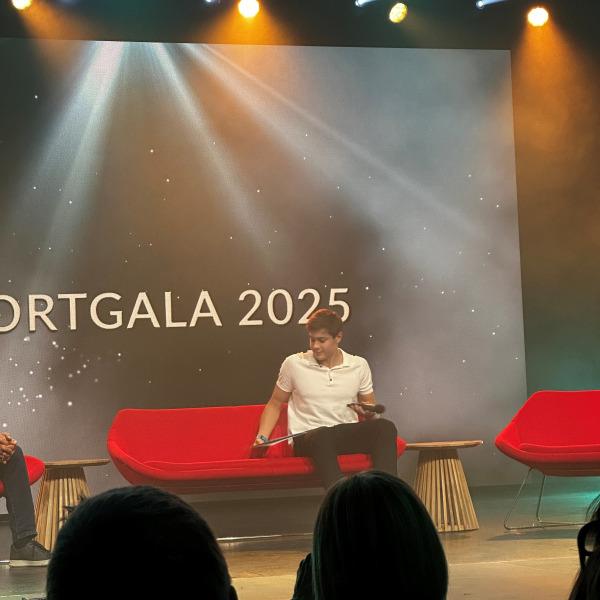 Mann in weißem Shirt sitzt auf rotem Sofa bei der Sportgala 2025, mit Lichtern und einer Präsentationsleinwand im Hintergrund.