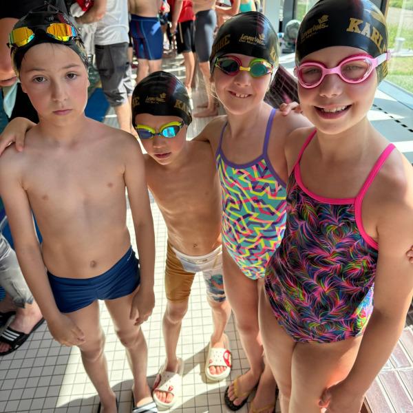 Vier Kinder in Badebekleidung und Schwimmkappen stehen zusammen am Poolrand, fröhlich lächelnd für ein Foto.