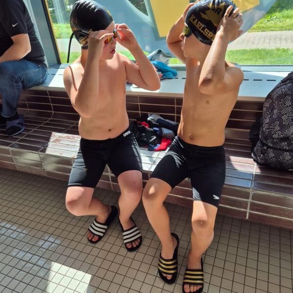 Zwei Jungen sitzen am Beckenrand, setzen Schwimmbrillen auf und tragen Badebekleidung und Badeschuhe.