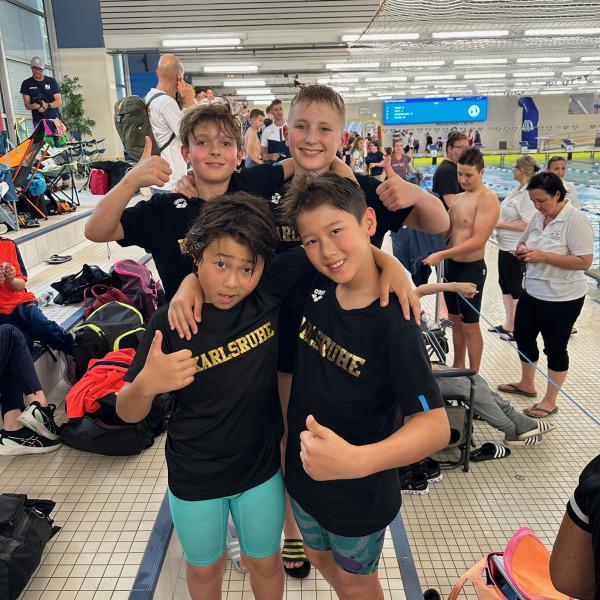 Vier Kinder in schwarz-gelben T-Shirts mit "Karlsruhe" posieren fröhlich am Beckenrand eines Schwimmbads.