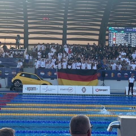 Zuschauer mit deutscher Fahne sitzen an einem Schwimmbecken während eines Wettkampfs, im Hintergrund ist eine Anzeigetafel sichtbar.