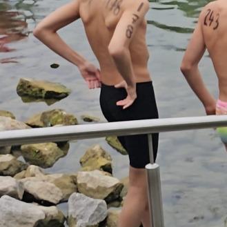 Zwei Schwimmer stehen am Ufer eines Gewässers, einer trägt eine Badekappe und hat Wettkampfnumbers auf den Armen.
