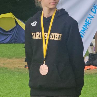 Mädchen in schwarzem Hoodie mit Medaille steht vor Zelt und Banner im Freien.