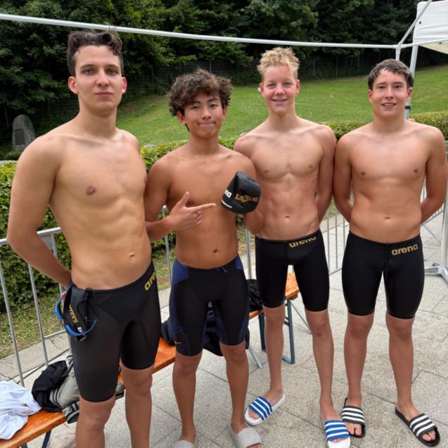 Vier junge Schwimmer stehen auf einer Freiluftanlage, tragen Badeanzüge und Halbmützen, im Hintergrund Bäume.