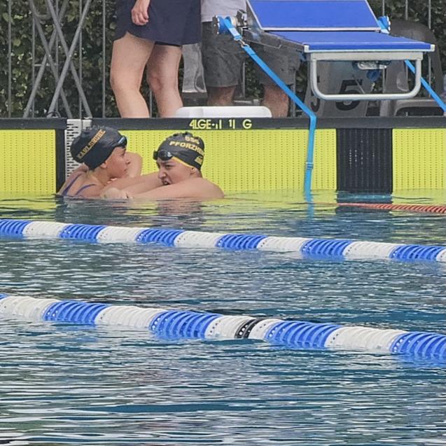 Zwei Schwimmerinnen in Badekappen umarmen sich im Wasser eines Schwimmbeckens, im Hintergrund stehen Zuschauer.
