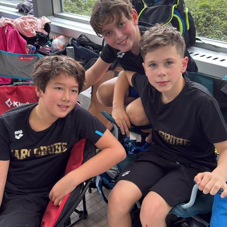 Drei Jungen in Sportkleidung sitzen lächelnd auf Campingstühlen in einer Sporteinrichtung.