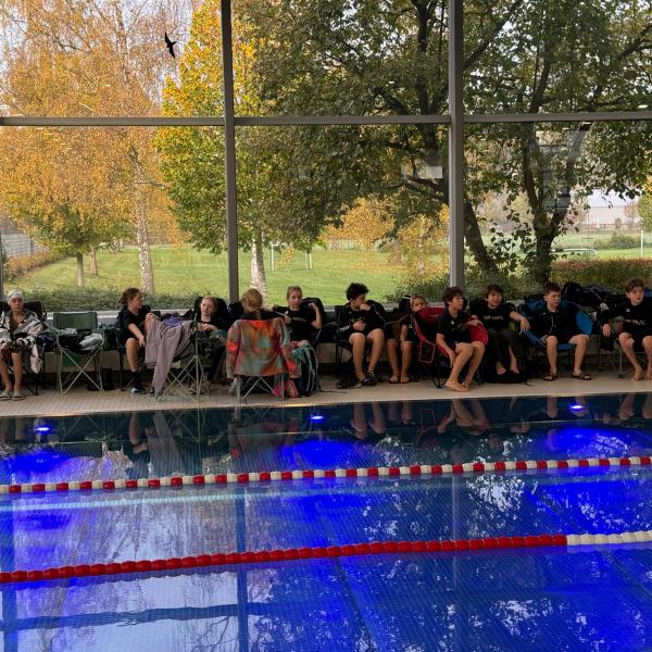 Gruppe von Schwimmern sitzt auf Stühlen am Beckenrand eines Pools, umgeben von herbstlicher Natur.