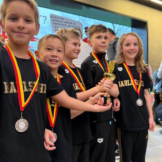 Gruppe von fünf Kindern in Sportkleidung mit Medaillen und einem Pokal in der Hand, lächelnd vor einem Hintergrund.