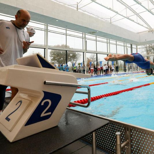 Schwimmer springt von Startblock 2 ins Wasser, während Trainer und Zuschauer am Beckenrand beobachten.