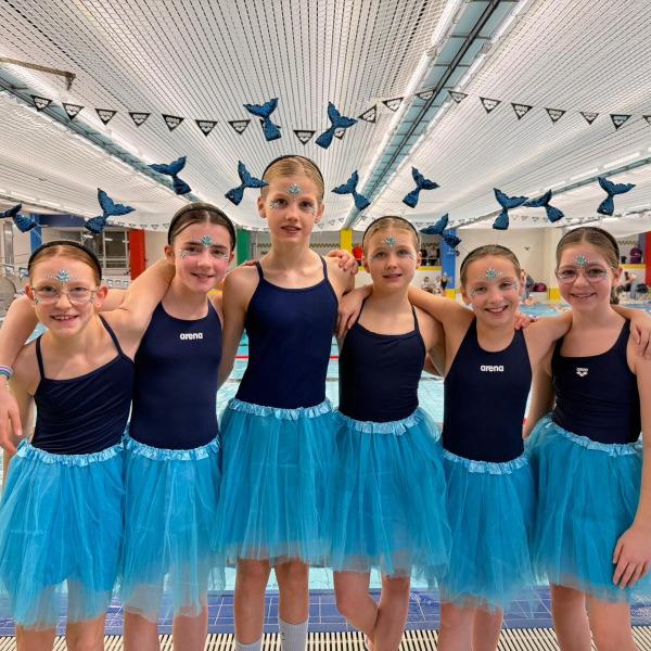 Sechs Mädchen im Schwimmanzug mit blauen Tüllröcken und Glitzer schminken sich am Schwimmbeckenrand.