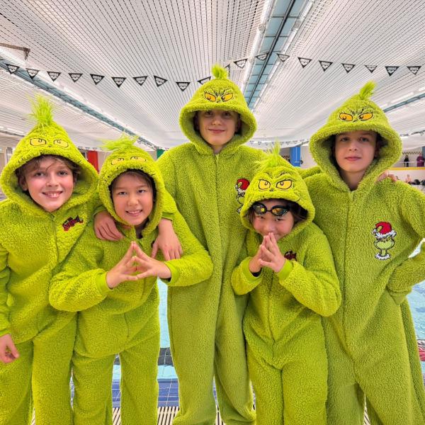Fünf Kinder in grünen Grinch-Mänteln posieren lächelnd am Beckenrand eines Schwimmbades.