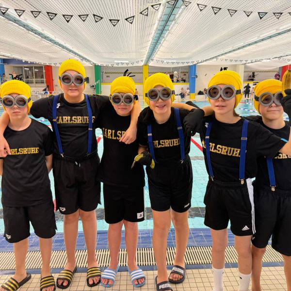 Sechs Kinder in Schwimmbekleidung mit gelben Mützen und Schwimmbrillen posieren am Beckenrand eines Schwimmbads.
