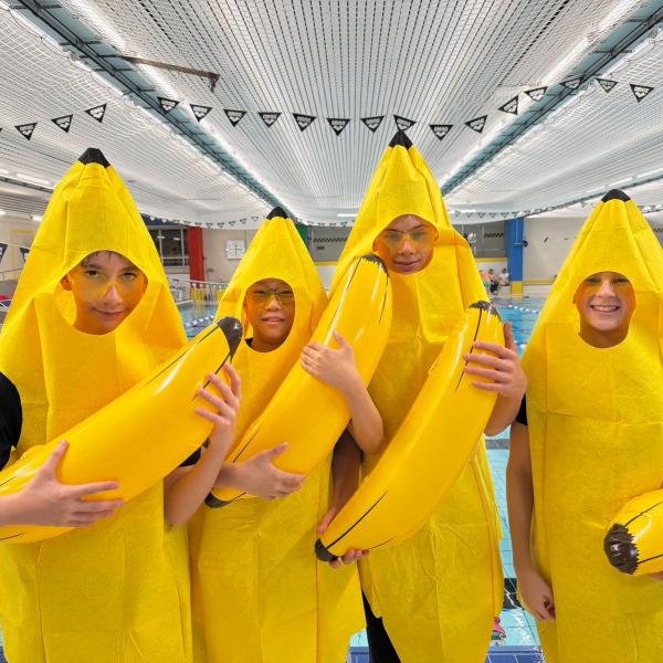 Vier Personen in Bananenkostümen stehen am Beckenrand eines Schwimmbads und halten gelbe Bananen in den Händen.