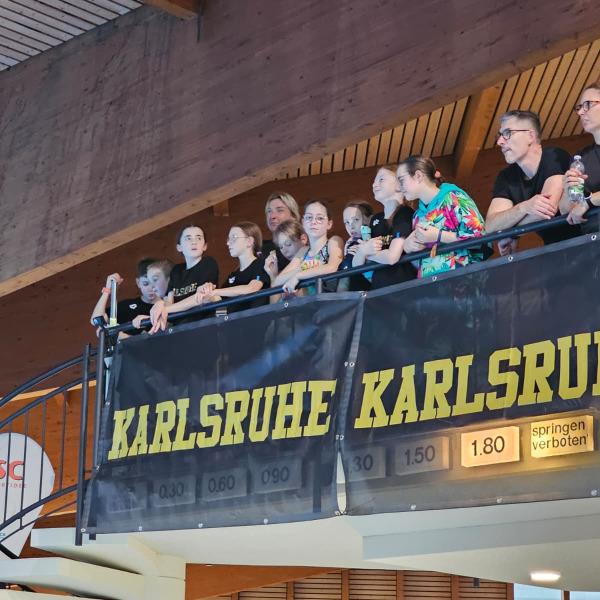 Junge Zuschauer stehen am Balkon einer Sporthalle mit einem Banner 'Karlsruhe'. Treppen im Hintergrund sichtbar.