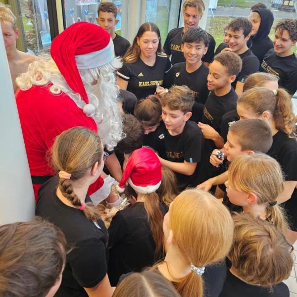 Santa Claus verteilt Geschenke an eine Gruppe von Jugendlichen in schwarzen T-Shirts, die ihm gespannt zusehen.