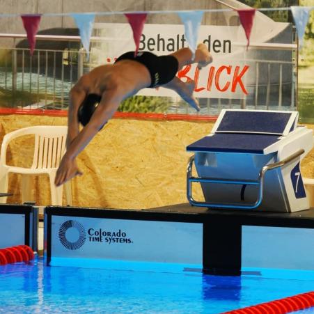 Schwimmer springt vom Startblock in ein Schwimmbecken, während ein Zuschauer im Hintergrund steht.