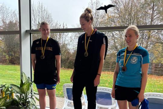 Drei Athletinnen mit Medaillen stehen auf dem Siegerpodest in einer Sporteinrichtung.