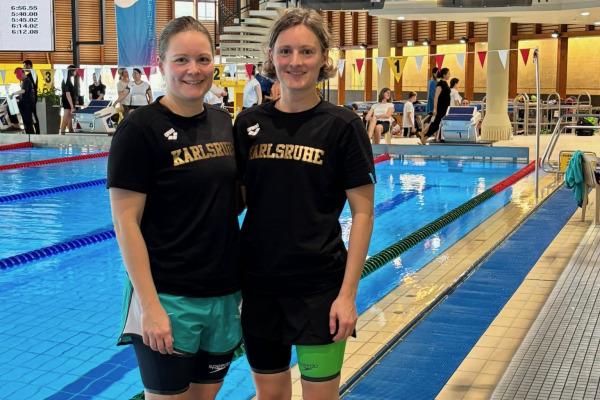 Zwei Schwimmerinnen in Sportbekleidung stehen am Beckenrand eines Schwimmbads mit Wettkampf-Atmosphäre.