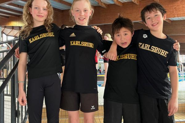 Vier Kinder in schwarzen T-Shirts mit der Aufschrift "Karlsruhe" stehen lachend auf einer Treppe in einem Schwimmbad.