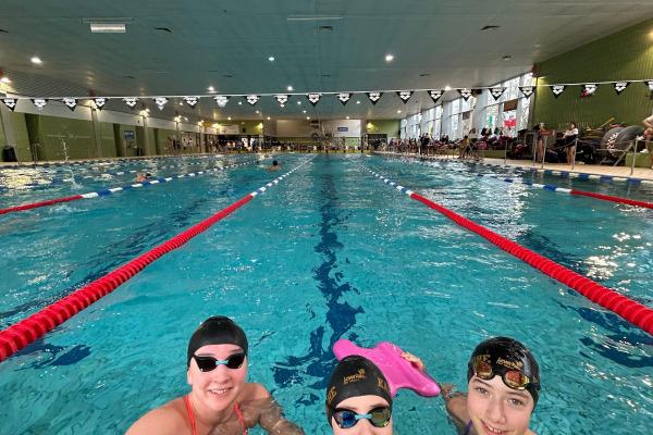 Drei schwimmende Kinder mit Schwimmbrettern posieren am Beckenrand eines Hallenbades.