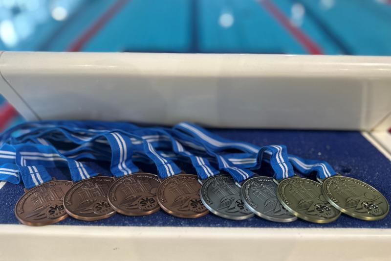 Acht Schwimmmedaillen in Bronze, Silber und Gold hängen an blauen Bändern auf einer Tischkante, dahinter ein Schwimmbecken.