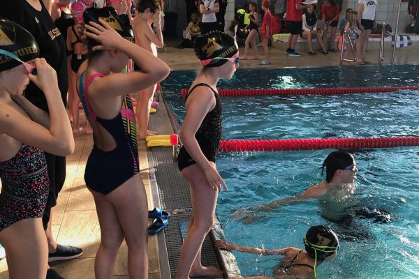 Kinder in Schwimmbekleidung stehen am Beckenrand, während im Wasser schwimmende Teilnehmer trainieren.